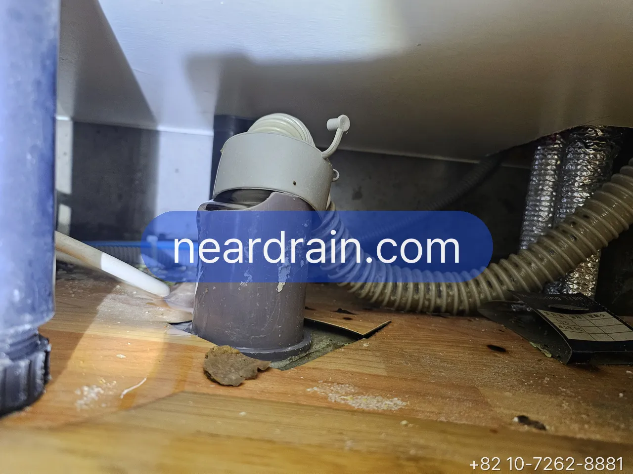 Grey corrugated drain hose pushed into a purple PVC floor stub under a cabinet, slime and grit at the joint on a wooden floor