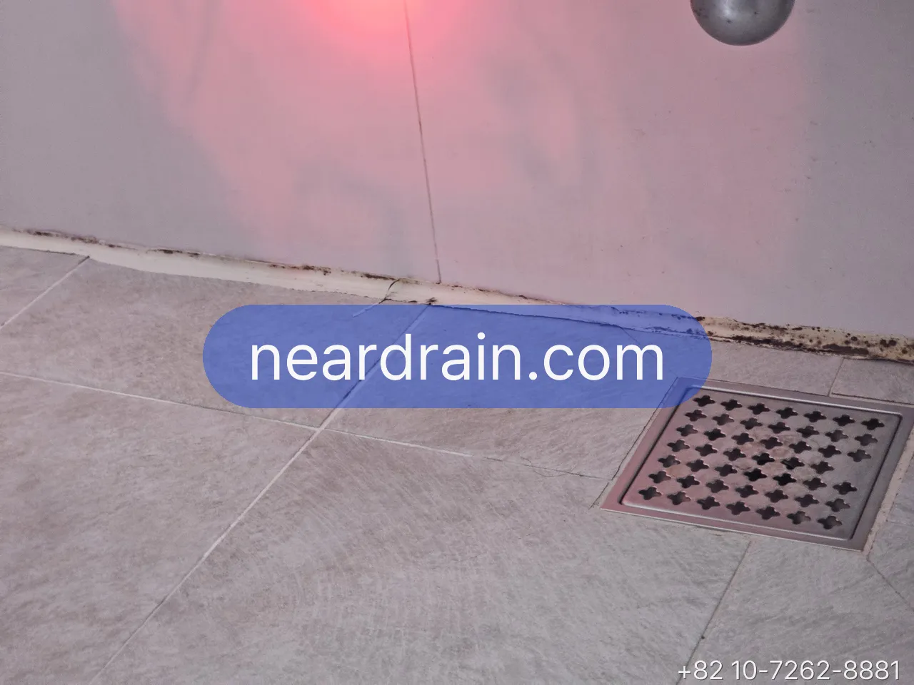 Square floor drain grate in a wet-room bathroom with water pooling around it and dark grout lines at the wall base