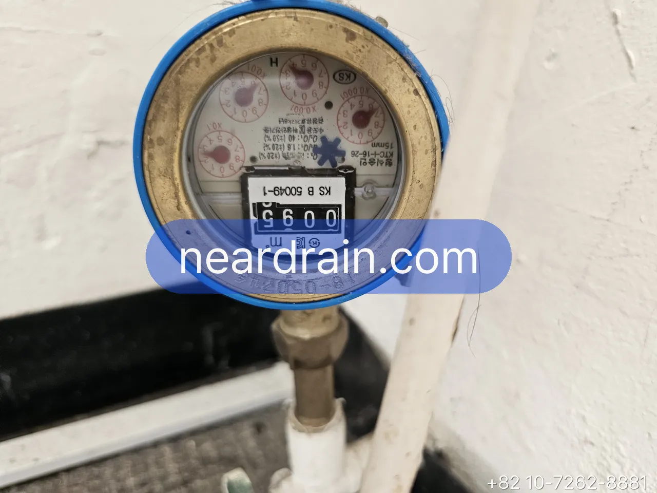 Water meter dial inside a Korean apartment meter box, blue-ringed meter on a vertical supply pipe with the shut-off below it
