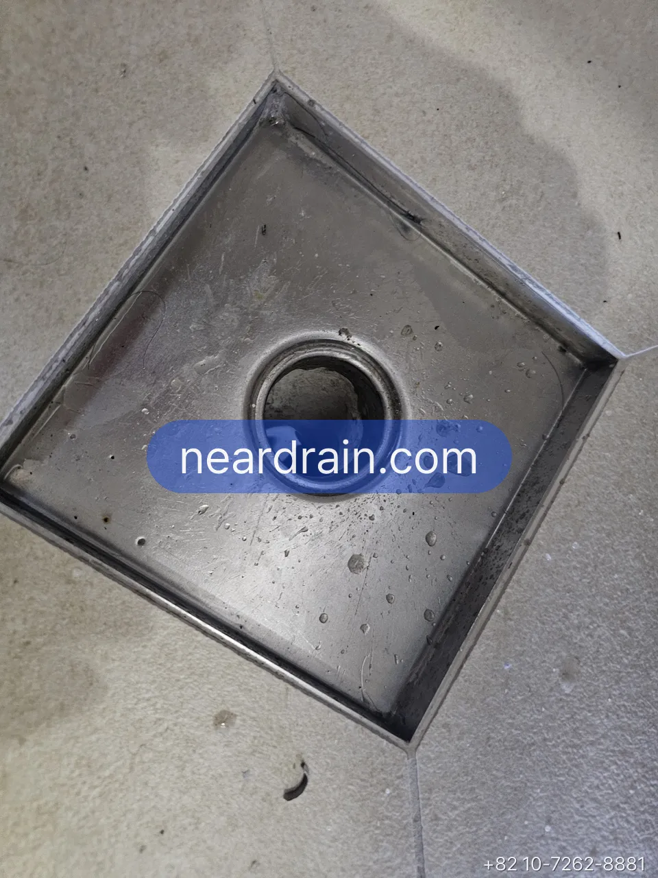 Square stainless balcony floor drain with the grate and cup removed, open pipe visible, water droplets on the steel