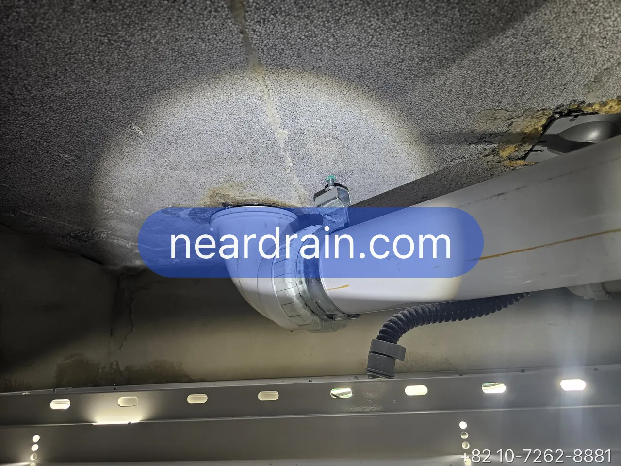 Ceiling in a downstairs unit lit by a torch, a drain pipe joint above the ceiling panel with brown water staining spreading from it