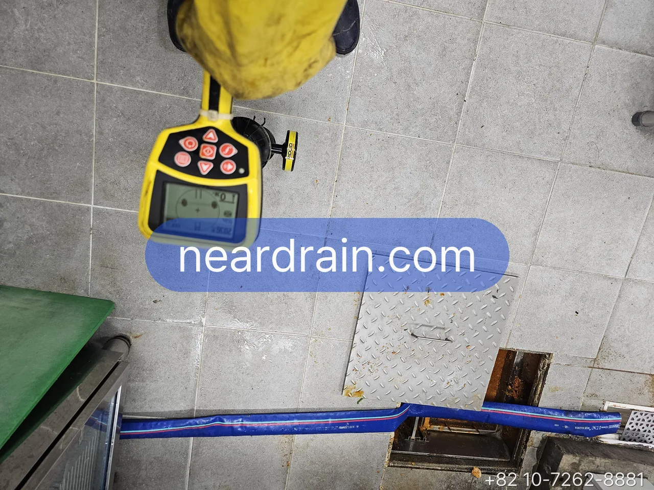 Acoustic leak locator with yellow sensor resting on a tiled floor beside an inspection hatch, blue supply hose crossing the tile