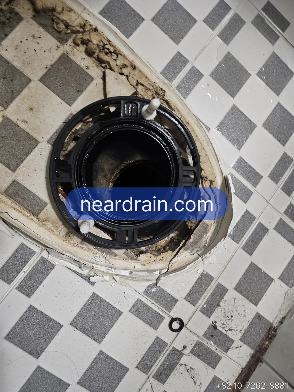 Toilet removed from a chequered-tile Korean bathroom floor, exposing the black floor flange and drain opening